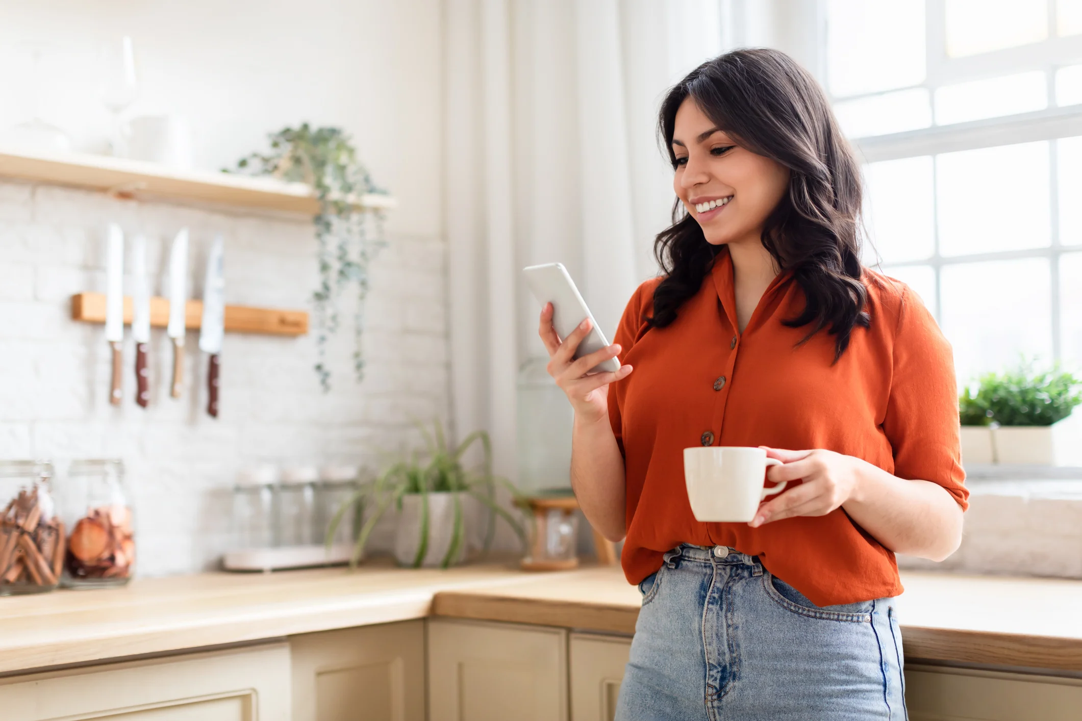 Persona sorridente che tiene una tazza mentre consulta uno smartphone in cucina.